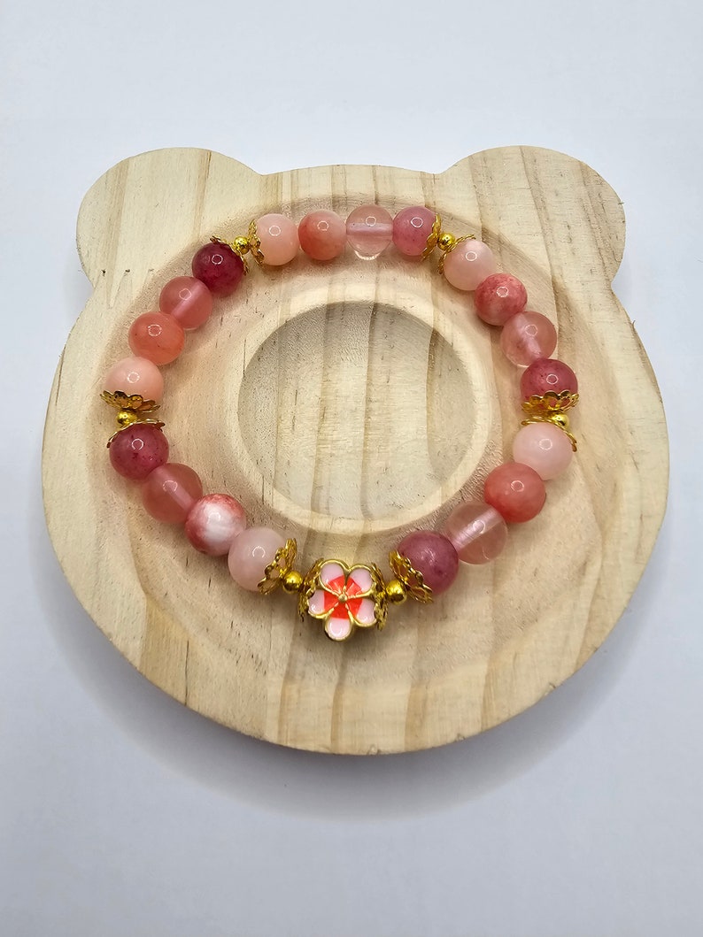 Crystal bracelet made of strawberry quartz, peach rice topaz, watermelon quartz and red chalcedony with gold flower and ball spacers and large gold sakura flower charm detailed with pink enamel.