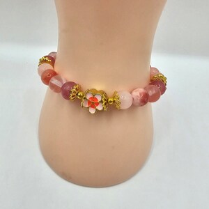 Crystal bracelet made of strawberry quartz, peach rice topaz, watermelon quartz and red chalcedony with gold flower and ball spacers and large gold sakura flower charm detailed with pink enamel.