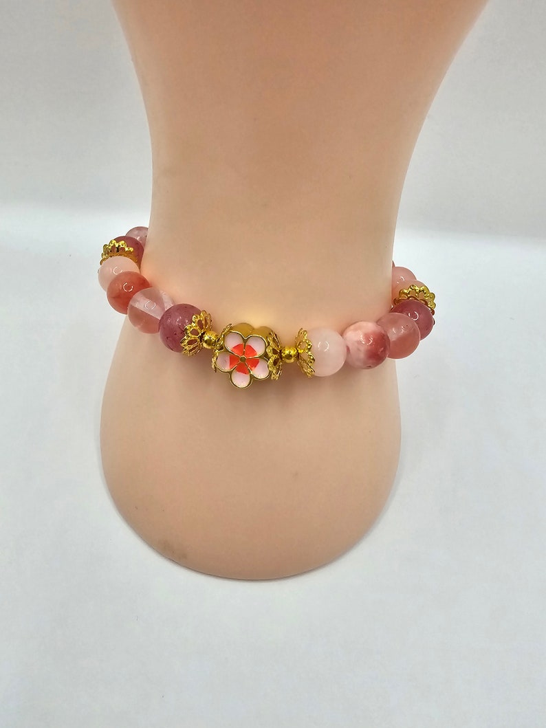 Crystal bracelet made of strawberry quartz, peach rice topaz, watermelon quartz and red chalcedony with gold flower and ball spacers and large gold sakura flower charm detailed with pink enamel.