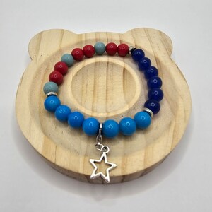 May include: A blue, red, and gray beaded bracelet with a silver star charm. The bracelet is on a wooden surface.