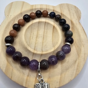 May include: A beaded bracelet featuring a variety of gemstone beads in shades of brown, purple, and blue. The bracelet has a silver charm of an open book. The bracelet is displayed on a wooden, bear-shaped coaster.