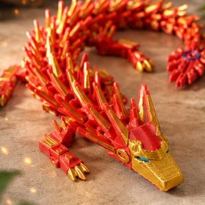 May include: A red and gold articulated dragon toy. The dragon has a detailed design with a red body and gold accents. The dragon is posed on a textured surface, showcasing its intricate features and design.