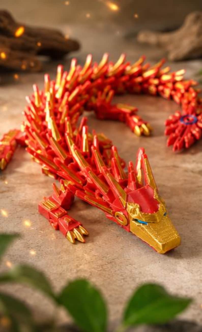 May include: A red and gold articulated dragon toy. The dragon has a detailed design with a red body and gold accents. The dragon is posed on a textured surface, showcasing its intricate features and design.