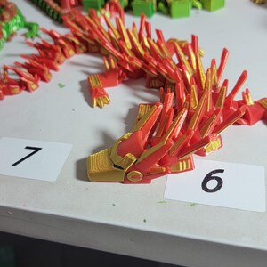 May include: A red and gold articulated dragon figurine. The dragon is made of plastic and has a detailed design. The head is gold with red accents. The dragon is on a white surface with number cards.