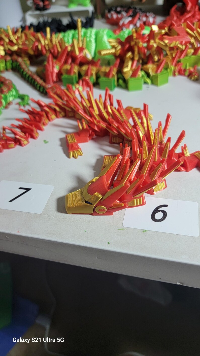 May include: A red and gold articulated dragon figurine. The dragon is made of plastic and has a detailed design. The head is gold with red accents. The dragon is on a white surface with number cards.