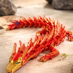 May include: A red and gold articulated dragon toy. The dragon features a detailed design with a red body and gold accents. The dragon's head and claws are gold. The dragon is posed on a textured surface.