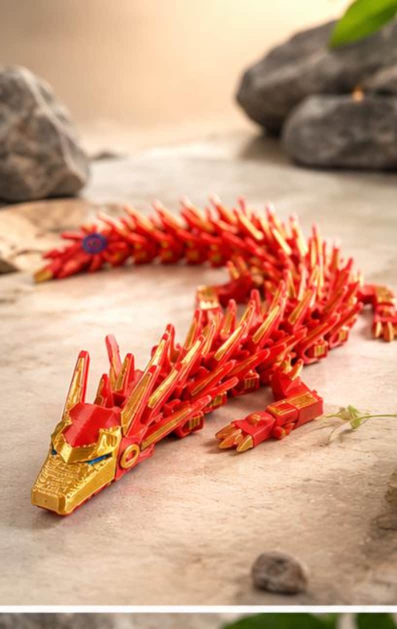 May include: A red and gold articulated dragon toy. The dragon features a detailed design with a red body and gold accents. The dragon's head and claws are gold. The dragon is posed on a textured surface.