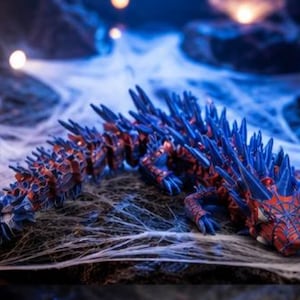 Pode incluir: Estatueta de dragão articulada em vermelho e azul, com um design de teia de aranha no rosto. O dragão está posicionado em uma superfície com teias de aranha falsas, criando um tema de Halloween ou fantasia.