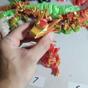 May include: A red and gold dragon figurine with intricate details, held in a hand. The dragon's head is gold with red accents, and the body is primarily red. Other 3D-printed figurines are visible in the background.
