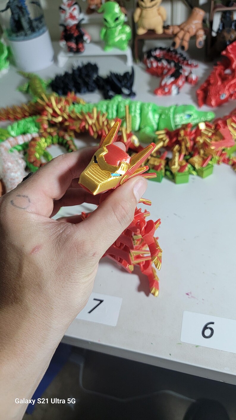 May include: A red and gold dragon figurine with intricate details, held in a hand. The dragon's head is gold with red accents, and the body is primarily red. Other 3D-printed figurines are visible in the background.