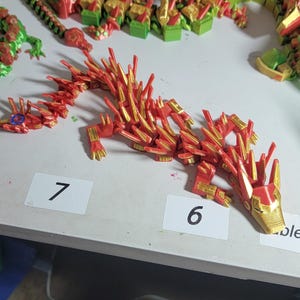 May include: A collection of 3D-printed articulated dragon toys in various colors. The primary dragon is red and gold, with a segmented body and spiky details. Other dragons are green, red, and black.
