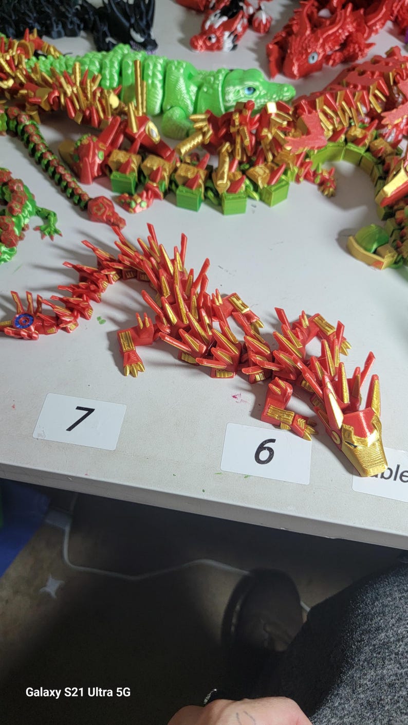 May include: A collection of 3D-printed articulated dragon toys in various colors. The primary dragon is red and gold, with a segmented body and spiky details. Other dragons are green, red, and black.