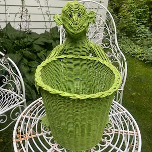 May include: A bright lime green wicker basket shaped like a monkey. The basket features a woven monkey head with dark, round eyes positioned above a deep, cylindrical woven container. The entire structure is crafted from painted wicker material.