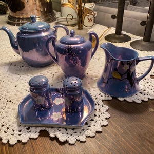 May include: A vintage blue tea set with a teapot, sugar bowl, creamer, and salt and pepper shakers. The set has a floral design and is displayed on a white lace doily. The set is on a wooden table.