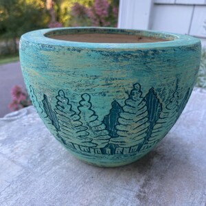 May include: A round ceramic planter with a distressed teal and light green finish. The exterior features a carved, dark green relief pattern depicting a dense forest of pine trees encircling the base of the bowl-shaped vessel.