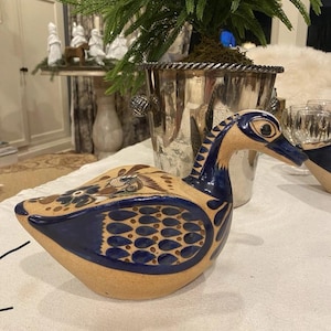 May include: A ceramic duck figurine with a tan body and blue accents. The duck has a long neck and beak, with detailed patterns on its wings and body. The figurine is displayed on a white surface.