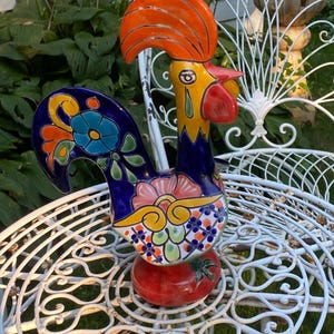 May include: A ceramic rooster figurine featuring a vibrant color palette of orange, yellow, red, and deep blue. The body is decorated with hand-painted floral and geometric patterns, including a prominent blue flower on the tail and a red base.