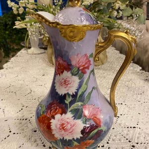 May include: A vintage porcelain pitcher with a lid, featuring a hand-painted floral design of pink and red carnations on a lavender background. The pitcher has gold accents on the handle and rim.
