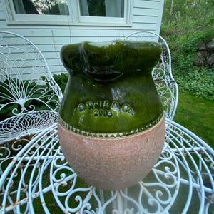 May include: A handcrafted ceramic pot with a green glazed top and a textured, light brown base. The pot features the text "Wolf & Co 2003" and sits on a white, ornate metal chair. The background includes a white house and green foliage.