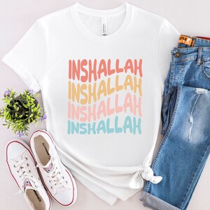 May include: White t-shirt with the phrase "Inshallah" repeated four times in a rainbow gradient. The text is in a retro font style.