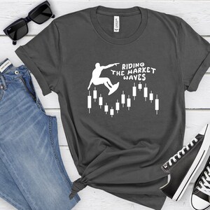 May include: A dark gray t-shirt with a white graphic of a person surfing on a candlestick chart. The text "Riding the Market Waves" is printed on the shirt.