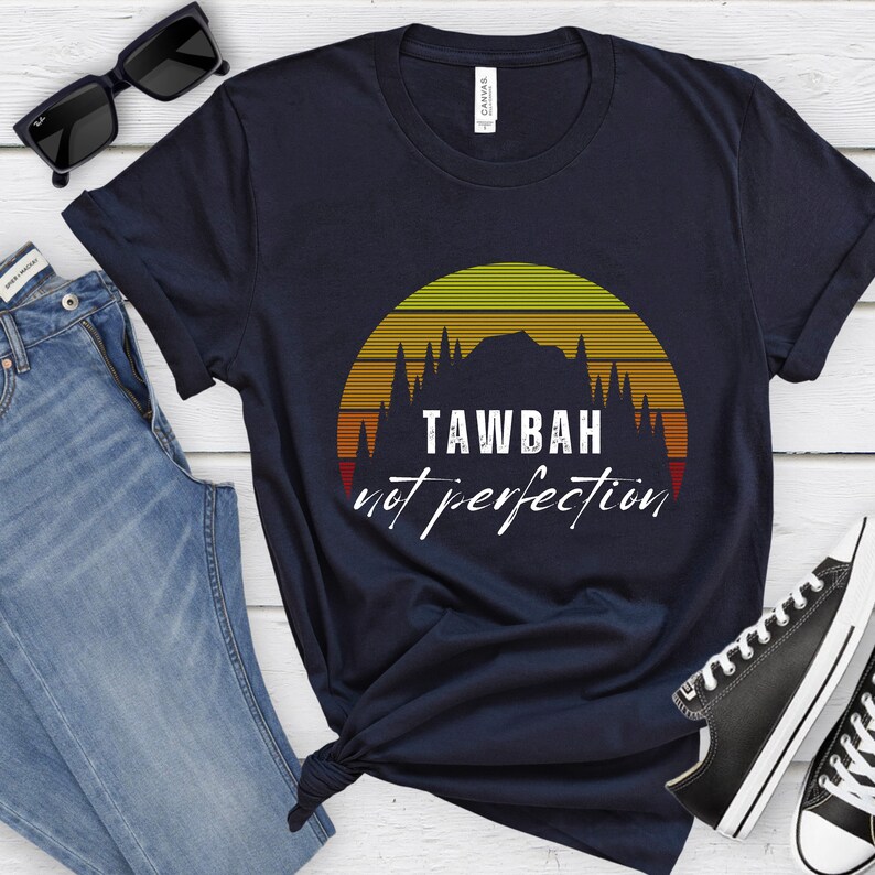 K&ouml;nnte beinhalten: Marineblaues T-Shirt mit einem Sonnenuntergangs-Design und dem wei&szlig;en Text "TAWBAH not perfection". Die Grafik zeigt eine Bergsilhouette und B&auml;ume. Das Shirt wird mit blauen Jeans, Sonnenbrille und Turnschuhen kombiniert.