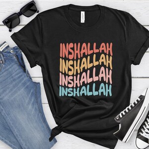May include: Black t-shirt with the phrase "Inshallah" repeated four times in a rainbow color gradient. The text is in a bold, stylized font.