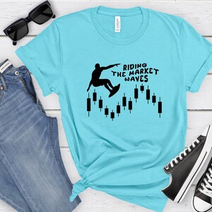 May include: A turquoise blue t-shirt with a black silhouette of a person surfing on a candlestick chart. The text "Riding the Market Waves" is printed on the shirt.