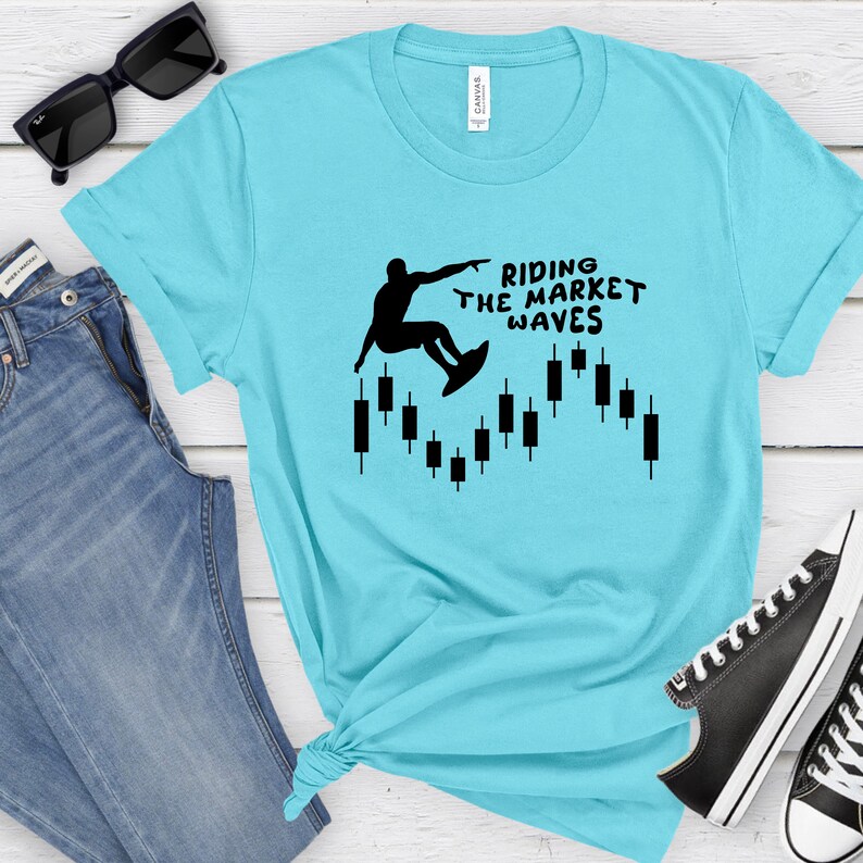 May include: A turquoise blue t-shirt with a black silhouette of a person surfing on a candlestick chart. The text "Riding the Market Waves" is printed on the shirt.