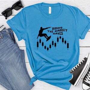 May include: A light blue t-shirt with a black silhouette of a person surfing on a candlestick chart. The text "Riding the Market Waves" is printed on the shirt.