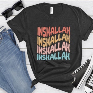 May include: A dark gray t-shirt with the word "Inshallah" repeated four times in a rainbow color pattern. The shirt is displayed on a white wooden background with a pair of jeans and sunglasses.