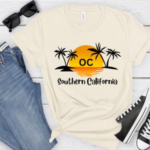 May include: A beige t-shirt with a black graphic of palm trees and a sunset with the text "OC Southern California".