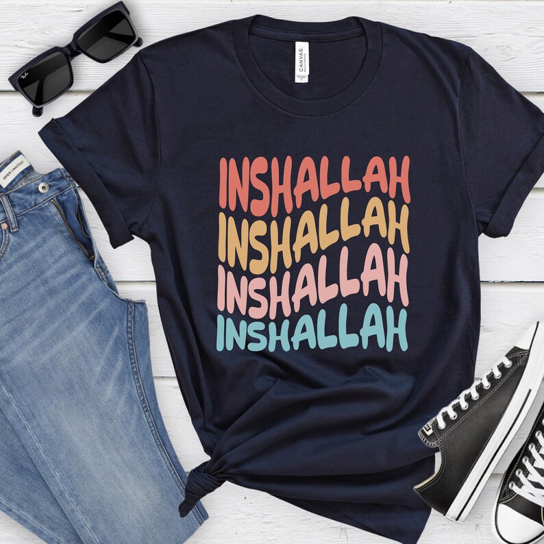 May include: A navy blue t-shirt with the word "Inshallah" repeated four times in a rainbow color gradient. The shirt is folded and displayed on a white wooden surface with a pair of sunglasses, a pair of jeans, and a pair of black sneakers.