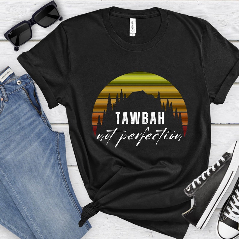 K&ouml;nnte beinhalten: Schwarzes T-Shirt mit einem Sonnenuntergang-Design und dem wei&szlig;en Text "TAWBAH not perfection". Das Design zeigt eine Bergsilhouette und B&auml;ume. Das Shirt wird mit blauen Jeans, Sonnenbrille und schwarzen Turnschuhen kombiniert.