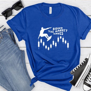 May include: A royal blue t-shirt with a white silhouette of a person surfing on a candlestick chart. The text "Riding the Market Waves" is printed on the shirt.