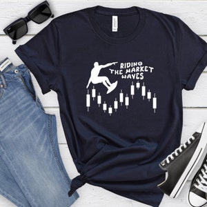May include: A navy blue t-shirt with a white graphic of a person surfing on a candlestick chart. The text "Riding the Market Waves" is printed on the shirt.