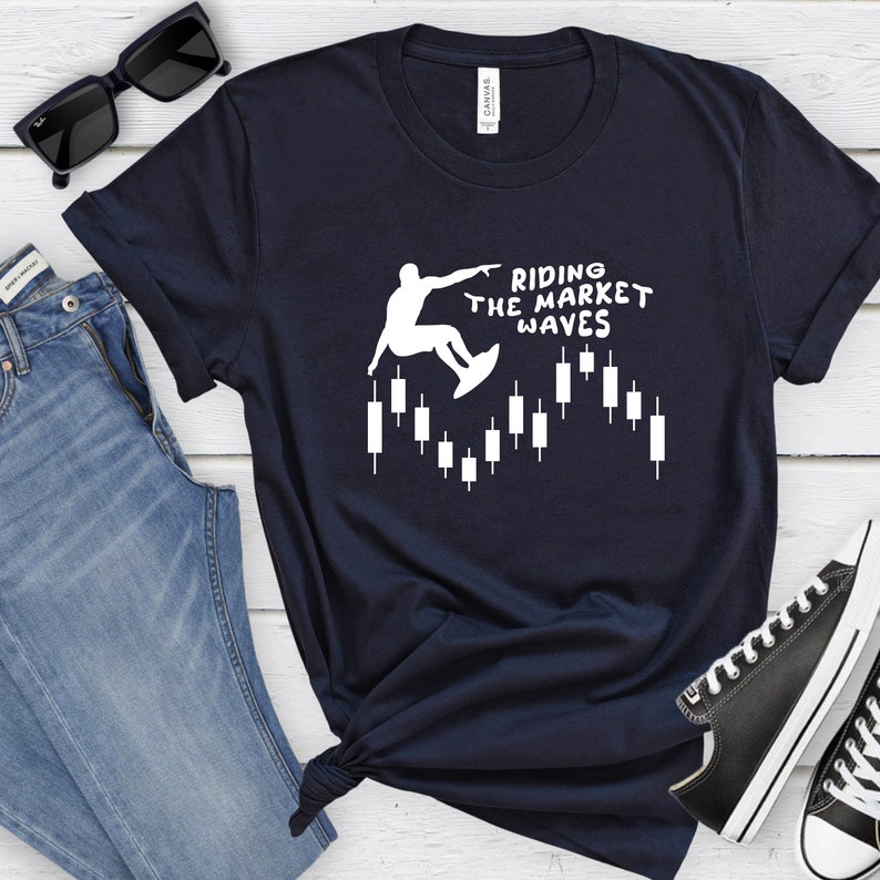 May include: A navy blue t-shirt with a white graphic of a person surfing on a candlestick chart. The text "Riding the Market Waves" is printed on the shirt.