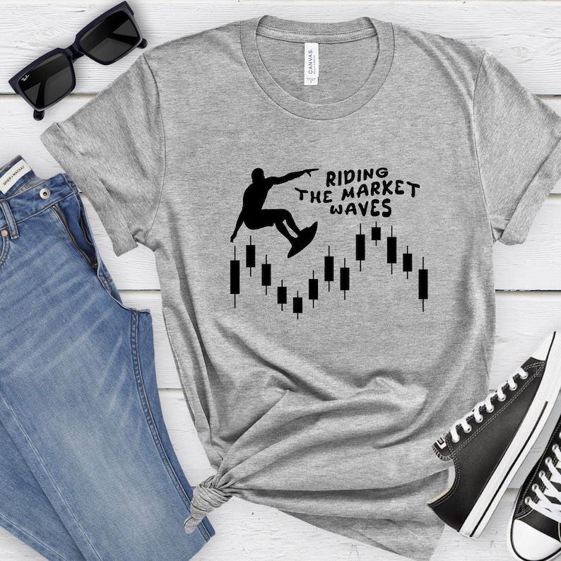 May include: A gray t-shirt with a black silhouette of a person surfing on a candlestick chart. The text "Riding the Market Waves" is printed on the shirt.