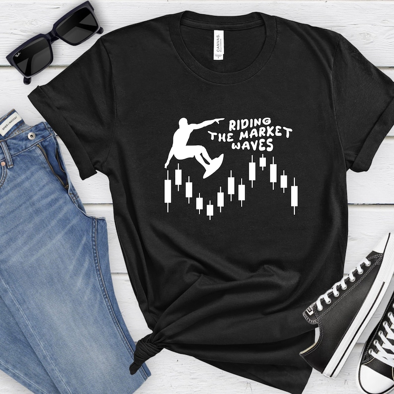 May include: Black t-shirt with a white graphic of a person surfing on a candlestick chart. The text "Riding the Market Waves" is above the graphic.
