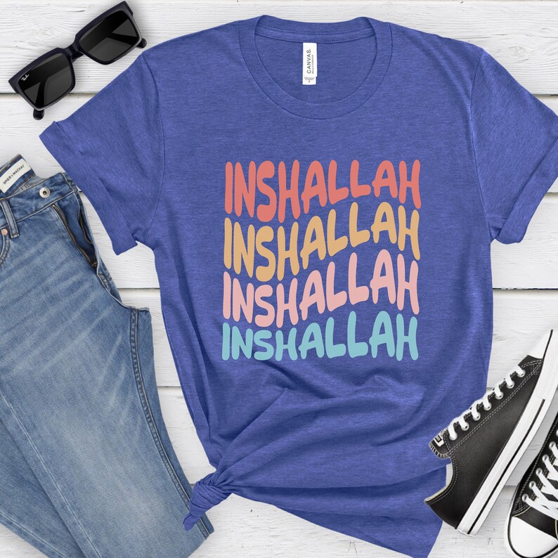 May include: A blue t-shirt with the word "Inshallah" repeated four times in a rainbow color pattern. The shirt is folded and displayed on a white wooden surface with a pair of jeans and black sneakers.