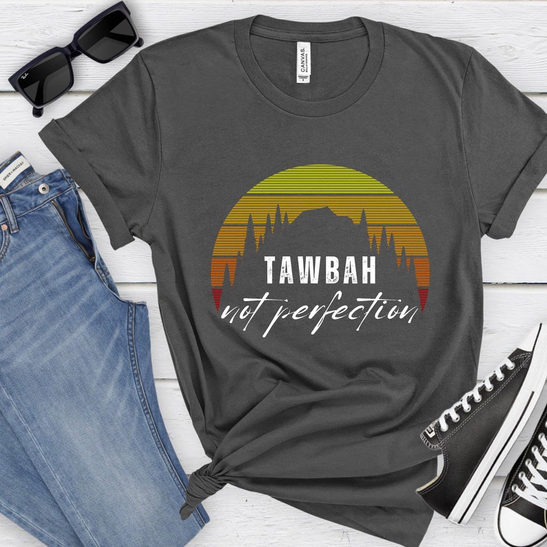 K&ouml;nnte beinhalten: Dunkelgraues T-Shirt mit einem Retro-Sonnenuntergangsdesign mit einer Bergsilhouette und dem Text "TAWBAH not perfection" in Wei&szlig;. Das Shirt wird mit blauen Jeans, Sonnenbrille und Turnschuhen pr&auml;sentiert.