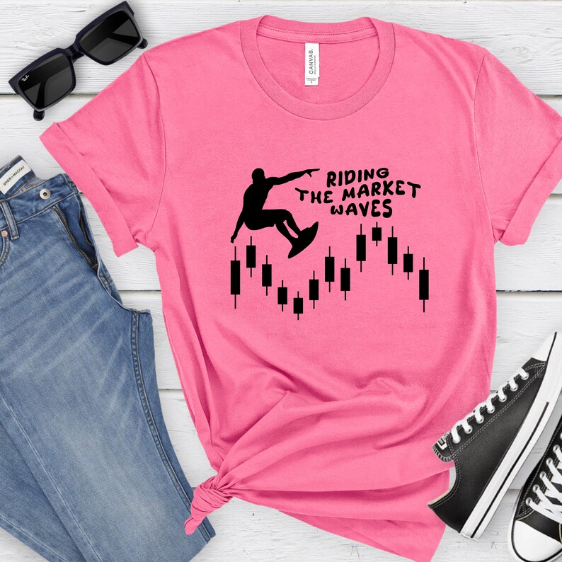 May include: Pink t-shirt with a black silhouette of a person surfing on a stock market chart. The text "Riding the Market Waves" is printed on the shirt.