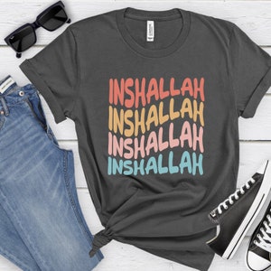 May include: A dark gray t-shirt with the word "Inshallah" repeated four times in a rainbow color scheme. The text is in a bold, handwritten style.