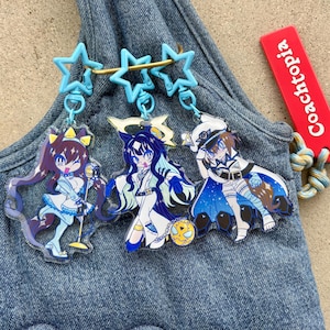 Könnte beinhalten: Drei Schlüsselanhänger mit Anime-Charakteren und blauen sternförmigen Anhängern. Die Charaktere haben lange Haare und tragen farbenfrohe Outfits. Ein Schlüsselanhänger hat ein rotes Etikett mit der Aufschrift "Coachtopia". Die Schlüsselanhänger befinden sich auf einem Jeans-Hintergrund.
