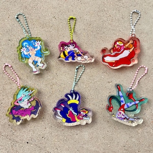 May include: Six colorful acrylic keychains with cartoon character designs. Each keychain features a different character in various poses, with vibrant colors like blue, red, and purple. The keychains have silver or gold-colored chain attachments.