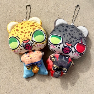 Puede incluir: Dos llaveros de peluche con cara de leopardo. Uno es amarillo con manchas marrones, el otro es gris con manchas negras. Ambos tienen grandes ojos verdes y bocas abiertas. El amarillo tiene un cuerpo azul y beige, el gris tiene un cuerpo rojo y negro.