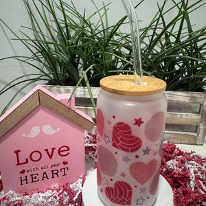 May include: A pink and white tumbler with a bamboo lid and a clear glass straw. The tumbler has a design of pink hearts with white stars and a red and white pattern. The tumbler is sitting on a white surface with pink and white tissue paper.
