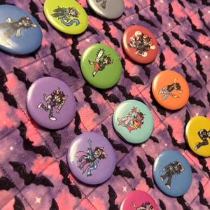 Könnte beinhalten: Eine Sammlung bunter runder Buttons mit Cartoon-Charakter-Designs. Die Buttons sind in verschiedenen Farben erhältlich, darunter Blau, Grün, Lila und Orange. Der Hintergrund ist ein lila Stoff mit einem Fledermausmuster.
