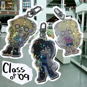 May include: Three keychains with cartoon character designs. The keychains feature holographic accents and silver-colored hardware. The characters have blonde, black, and light blonde hair, wearing various outfits. The text "Class of '09" is visible on a piece of paper.