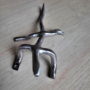 May include: A silver metal sculpture of a stylized bird with a long neck and a curved beak. The sculpture has a three-pronged base.